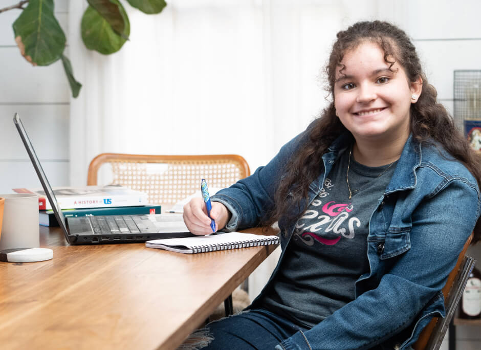 Estudiante sonriendo a la camara, sentada en su mesa con la imagen de su portatil